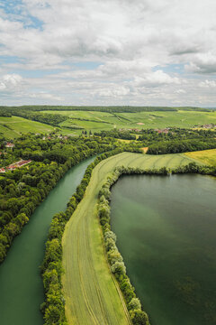 Aerial view of the Marne river and adjacent lake surrounded by lush green fields, dense trees, and rolling hills under a cloudy sky in Barzy-sur-Marne, Hauts-de-France, France.