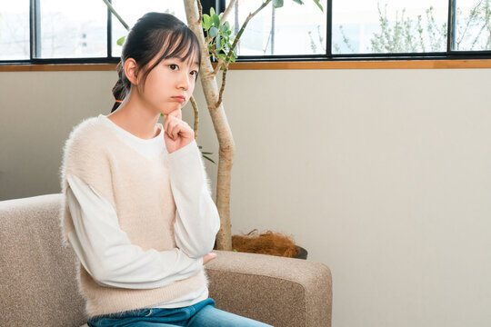 A young girl, either an elementary or middle school student, sits on the living room sofa, thinking and worrying.