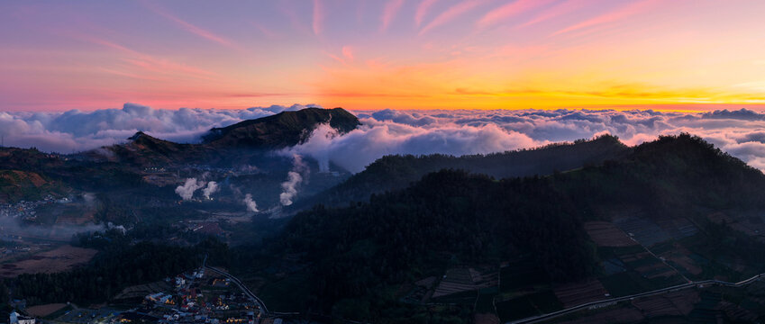 Aerial view of Dieng Plateau with volcanic steam rising from fumaroles amidst lush mountains and agricultural fields under a vibrant sunrise sky in Indonesia.