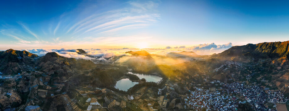 Aerial view of Dieng Plateau featuring Telaga Warna lake, terraced fields, and a village nestled among volcanic mountains at sunrise with golden light rays in Indonesia.