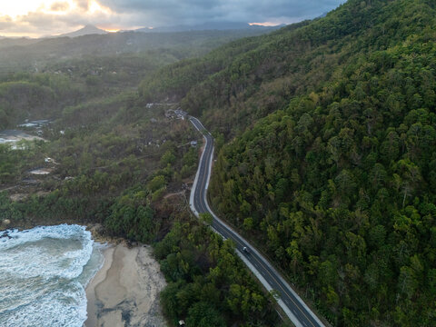 Aerial view of a winding coastal road surrounded by lush green hills and dense forest next to the ocean at sunset in Pacitan, East Java, Indonesia.