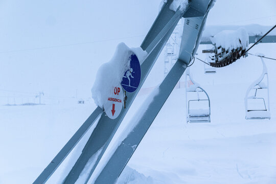 Close-up of a metal ski chairlift structure and a blue and red emergency stop sign deeply covered in fresh snow during a winter storm at a ski resort.