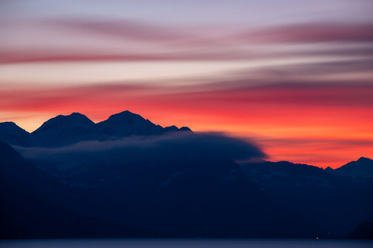 View of the dramatic sunset over the snow-capped peaks of the Bernese Alps with low-hanging clouds and the calm waters of Lake Thun in Interlaken, Canton of Bern, Switzerland.