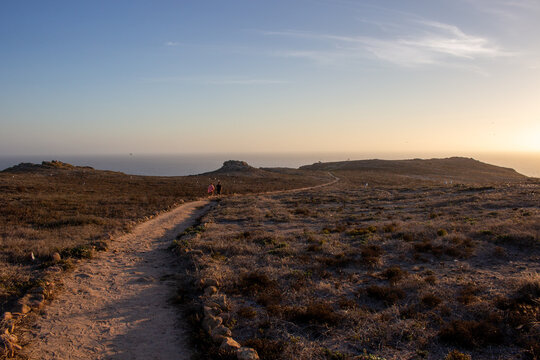 View of a dirt path winding through the dry, rocky landscape of the Berlengas archipelago with two people walking towards the ocean at sunset in Leiria, Portugal.