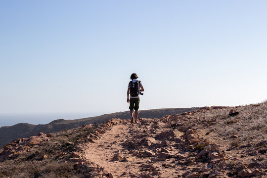 View of a hiker with a backpack walking along a rocky dirt path on the Berlengas islands with the ocean in the background in Leiria, Portugal.