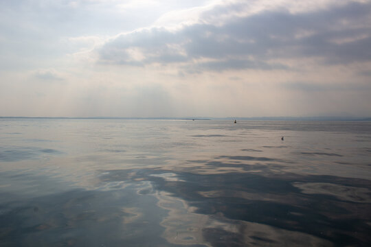 View of Garda Lake with calm water and soft light filtering through clouds on the horizon in Lombardia, Italy.
