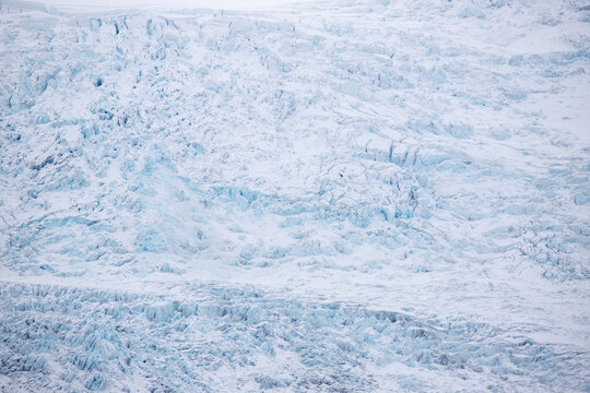 View of the textured surface of a glacier with deep blue ice, jagged crevasses, and layers of snow in Iceland.