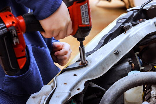 Car worker works on car engine with power drill in garage engine is open tools are around.