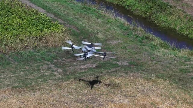 High angle view of a large hexacopter drone flying low over a green and yellow agricultural field, inspecting crops and casting a shadow on the ground as it moves across the landscape