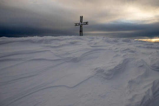 View of the summit cross on the snow-covered peak of the Apuan Alps with wind-swept snow patterns under a dramatic cloudy sky in Minucciano, Tuscany, Italy.