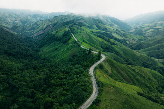 Aerial view of a winding asphalt road cutting through lush green mountains and tropical rainforest.