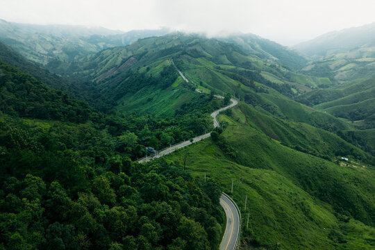 Aerial view of a winding asphalt road cutting through lush green mountains and tropical rainforest.