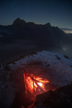 View of a glowing campfire on a snow-covered mountain peak under a starry night sky with the Apuan Alps in the background in Minucciano, Tuscany, Italy.
