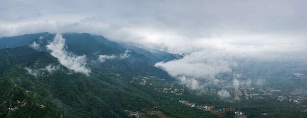 A panoramic view of forested mountains with drifting clouds and scattered settlements in the valley below under an overcast sky. © tai