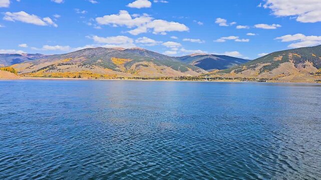 Boat level view across a calm mountain lake, with blue water, golden autumn hills, scattered aspens, evergreen forest, and distant peaks beneath bright clouds on a sunny day.