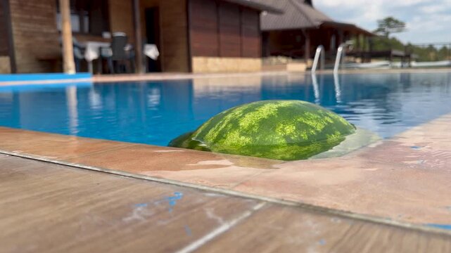 A striped watermelon bobs against the edge of a blue swimming pool in close up, with shallow focus revealing wet tan tiles, a metal ladder, and a wooden lodge beyond the water.