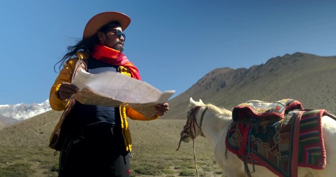 Annapurna Circuit, Mustang, Nepal, low-angle shot of a male traveler wearing a cowboy hat and sunglasses holding open a paper map next to a saddled horse