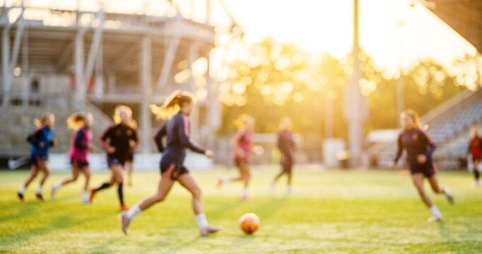 A dynamic soccer practice session with players running and kicking a ball on a sunny field. The blurred background features a stadium and trees.