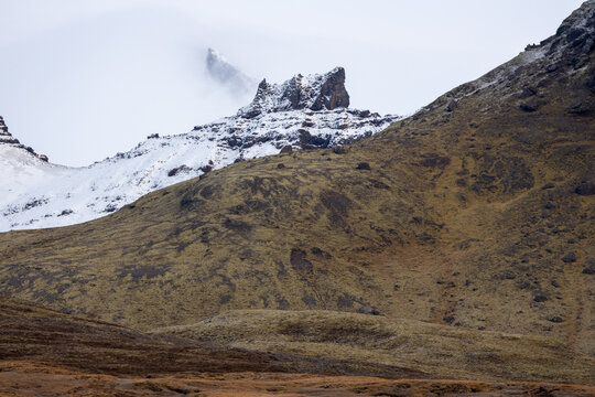View of a snow-capped mountain peak partially shrouded in clouds above mossy brown slopes in Iceland.