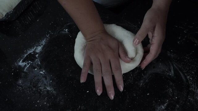 Close up of hands shaping and kneading a piece of homemade dough on a dark floured work surface.