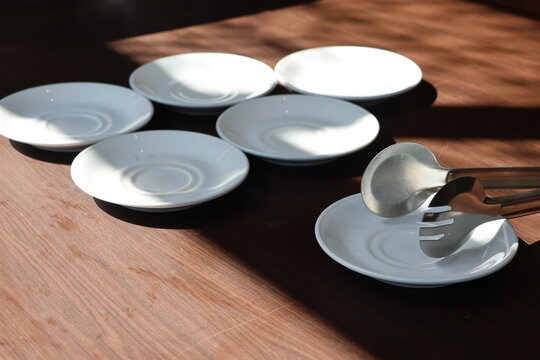 Sunlight casts soft shadows across minimalist tableware. Five clear plates rest beside metal tongs. Neutral tones evoke calm, quiet dining moments. Ideal for design, lifestyle, or editorial projects