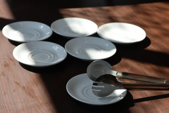 Sunlight casts soft shadows across minimalist tableware. Five clear plates rest beside metal tongs. Neutral tones evoke calm, quiet dining moments. Ideal for design, lifestyle, or editorial projects