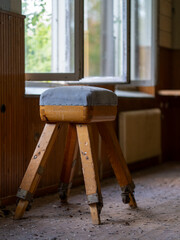 Old wooden gymnastics vaulting stool with a padded top standing on a dusty floor inside an abandoned rural school building. © HelenaUpNorth