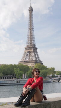 Solo traveler Asian woman in a red cardigan and black boots sits on a ledge overlooking the river Seine with a blurred Eiffel Tower in the distance