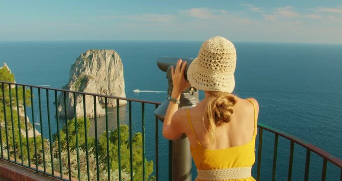 Woman tourist using telescope at panoramic seaside cliff lookout point with Faraglioni sea stacks in Capri