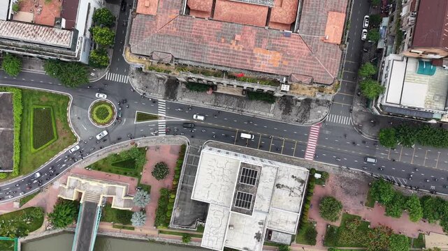 Top down aerial tracking across a district, passing a small planted roundabout, a road underpass and a large tiled roof building among the shophouses, Ho Chi Minh City, Vietnam.