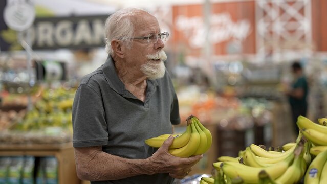 An elderly man is in a grocery store holding a bunch of bananas and looking at other items around him while shopping in the daylight
