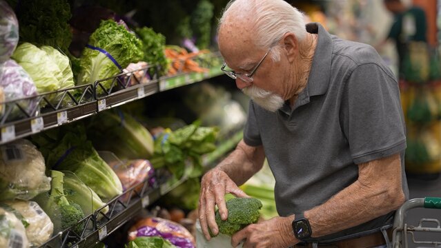 An elderly man examines broccoli while shopping for vegetables in a grocery store during the morning hours
