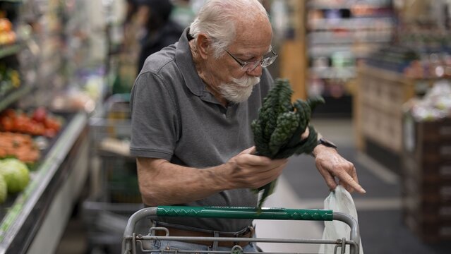An elderly man is shopping in a grocery store. He holds a vegetable while looking at his cart filled with items