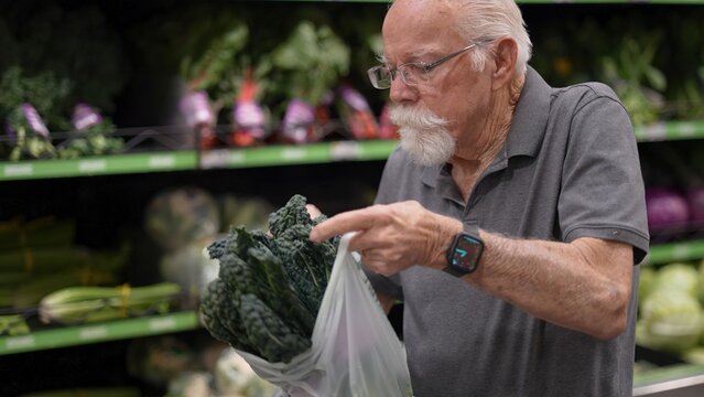An elderly man stands in a grocery store looking at vegetables inside a bag. He is selecting fresh produce for his shopping