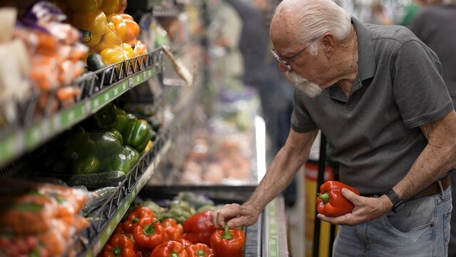 An elderly man browses through red peppers in a grocery store aisle while shopping for fresh produce