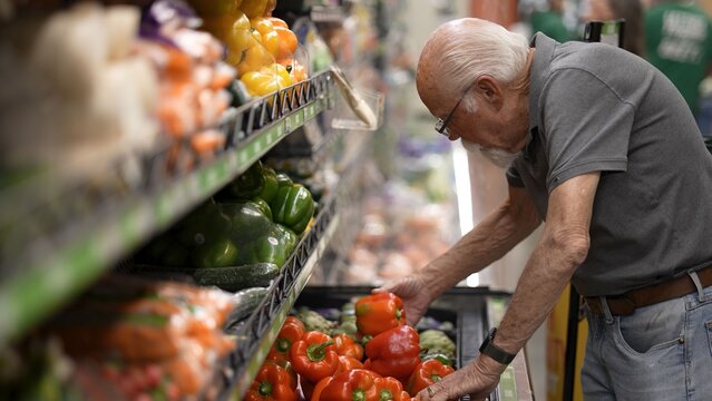 An elderly man shops for fresh vegetables in a grocery store. He carefully examines bell peppers on the shelf