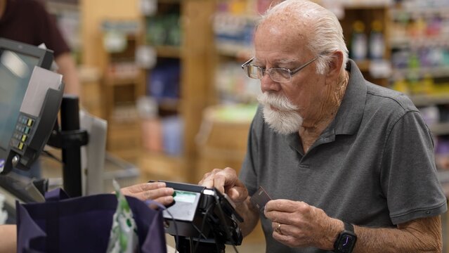 An elderly man is at the grocery store using his card to pay for his items at the checkout. He seems focused on the process
