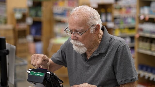 An elderly man is shopping at a grocery store, using a card to check out his items. He focuses on completing his purchase