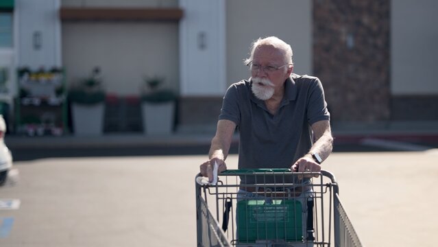 An elderly man is at a grocery store, pushing a cart as he shops for items. He is focused on his task in the parking lot