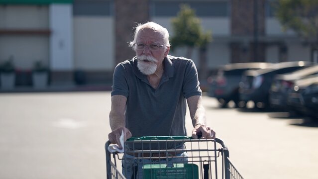 An elderly man shops in a grocery store parking lot, using a shopping cart to carry his items as he walks through the area