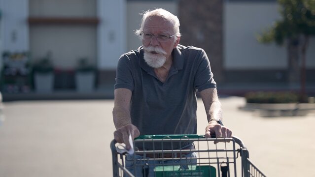 An elderly man is shopping at a grocery store parking lot with a cart. He seems focused on his task while enjoying the outdoor atmosphere