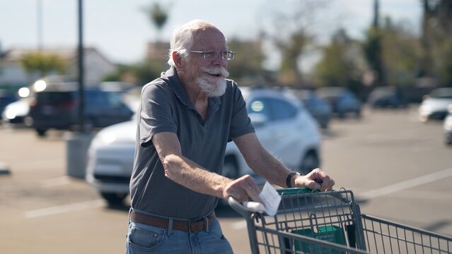 An elderly man shops for groceries in a parking lot while pushing a cart filled with items. He wears glasses and has a beard