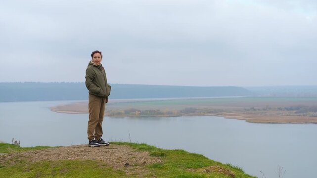 Woman standing on cliff contemplating river landscape. Full-length view of a pensive woman standing on the edge of a grassy cliff, contemplating the scenic river view on a cloudy, overcast day