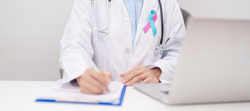 doctor using laptop and report notes on notebook. blue and pink ribbon concept of October male Breast Cancer Awareness month, National Cancer Survivors Month and Pink October Healthcare