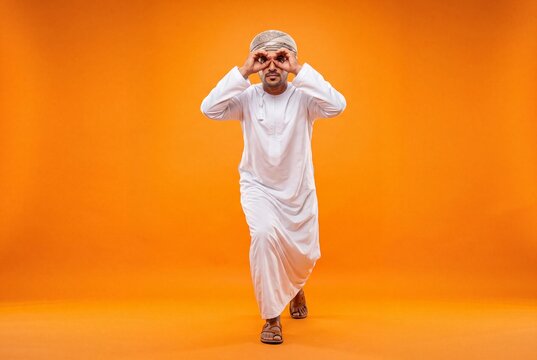 Omani Man in Traditional Dishdasha Making Binocular Gesture on Orange Studio Background