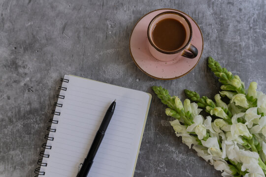 Coffee, notebook, and flowers rest on a textured gray surface. Black pen lies beside open lined pages, ready for notes. Soft pink cup holds warm beverage, adding calm to the scene