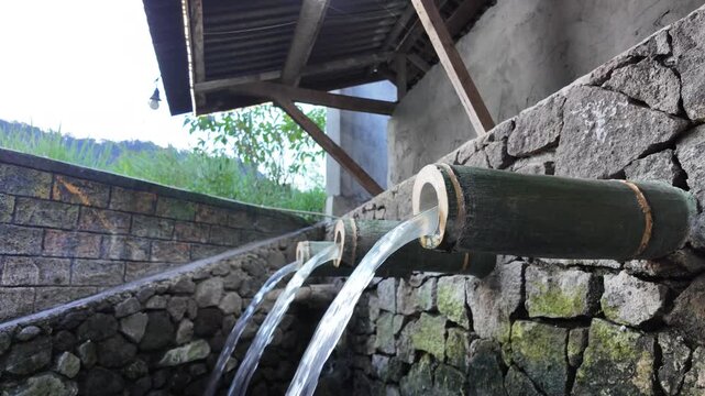 Fresh mountain water pours steadily from a traditional bamboo pipe over an old stone wall, creating a peaceful scene that reflects the simple beauty of rural life.