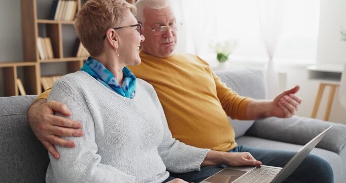 Seniors couple video chat online on laptop home. They sit together on a sofa using technology to connect with family or telehealth in a home setting. Clear concept of digital communication.