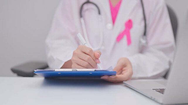 Professional doctor wearing lab coat using laptop and taking report notes on notebook for Breast Cancer Awareness month, World Cancer Day, National Cancer Survivors Month and Pink October in hospital