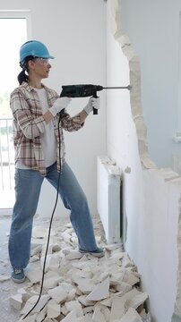 Female construction worker demolishing a wall with a hammer drill, wearing protective gear, generating rubble pile during renovation project